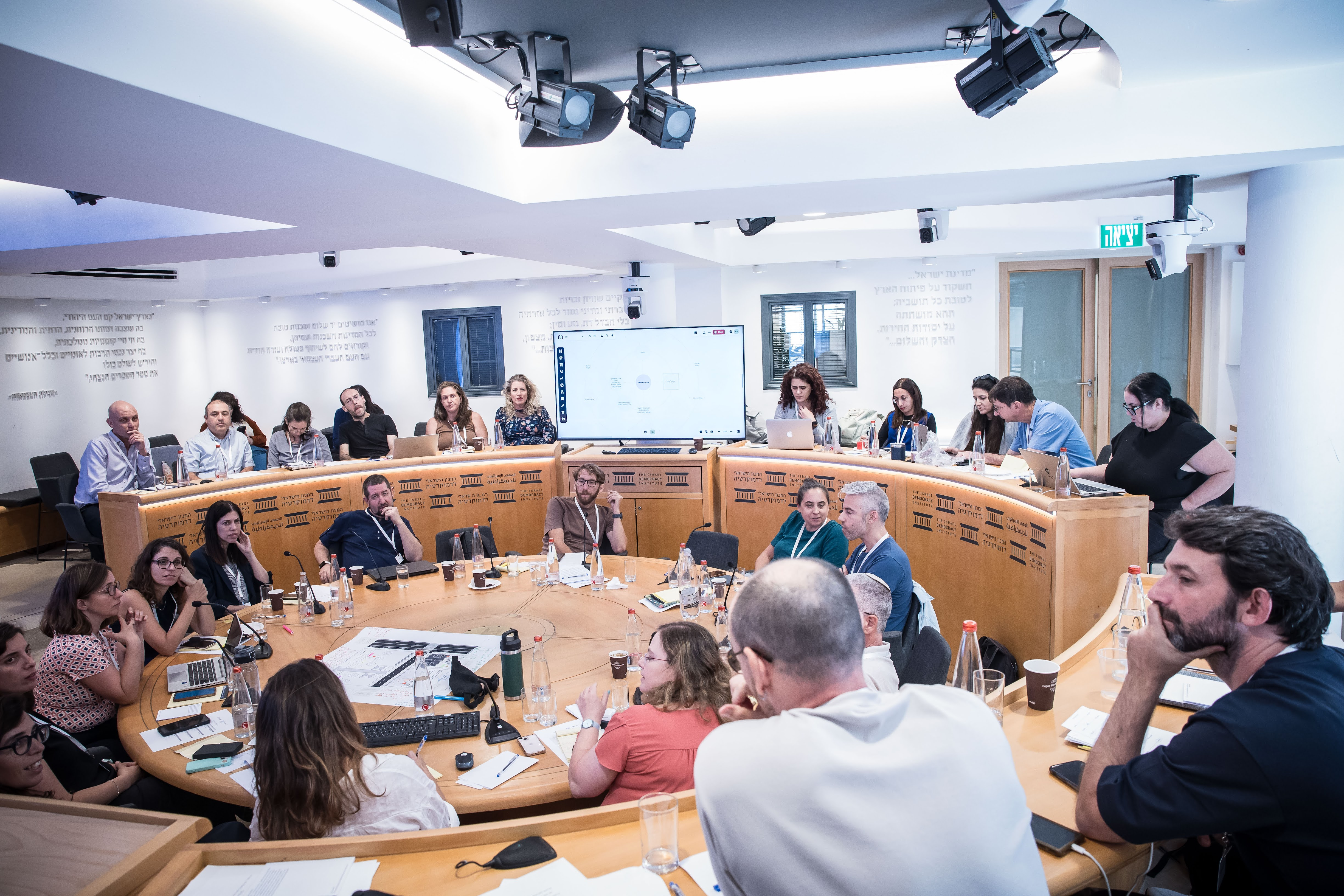 People gathered in a modern conference room for a collaborative meeting, seated around a circular table with laptops and notes, while a presentation is displayed on a large screen.