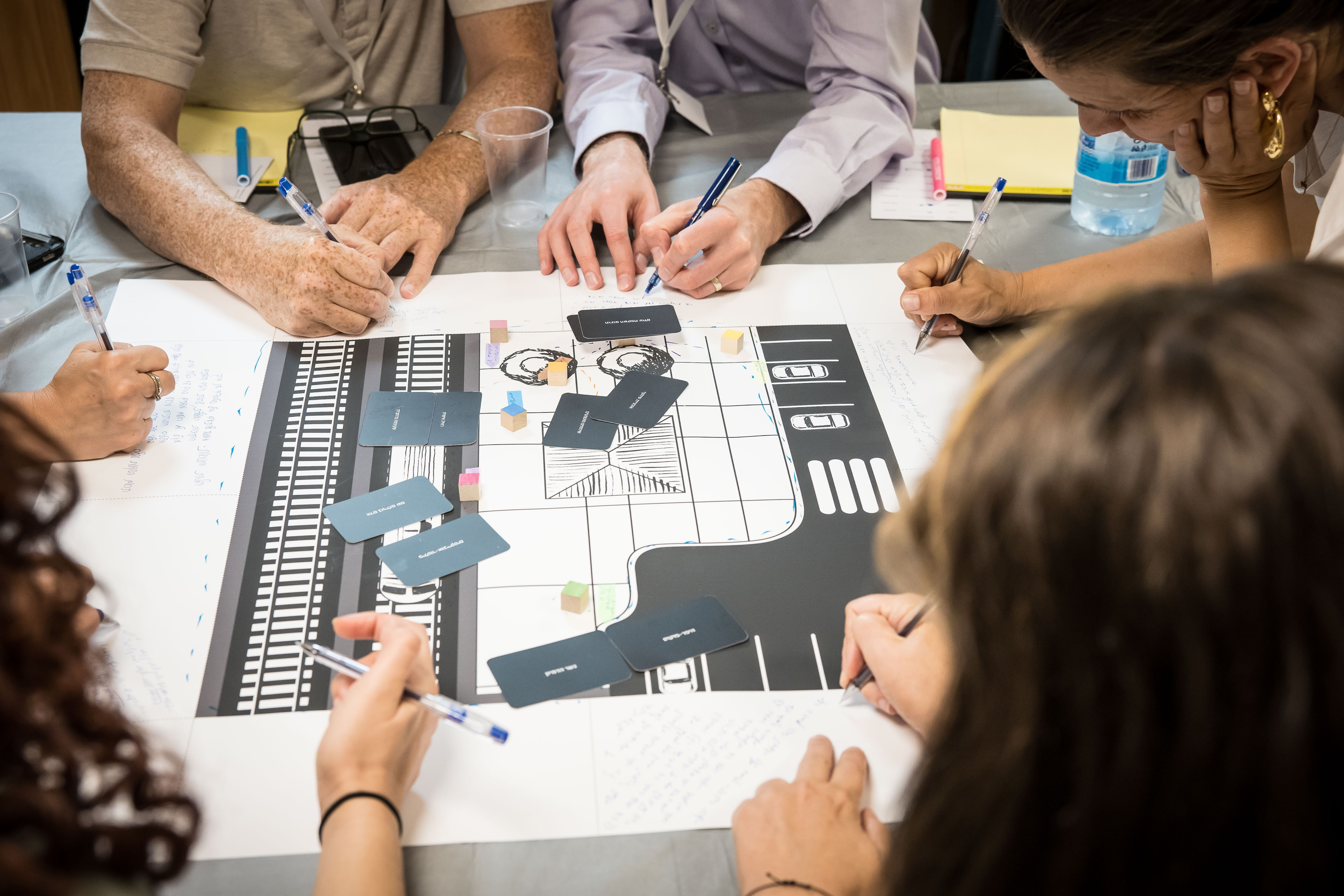 People collaborating around a table, writing notes and interacting with a printed map covered with vocabulary cards and small cubes.